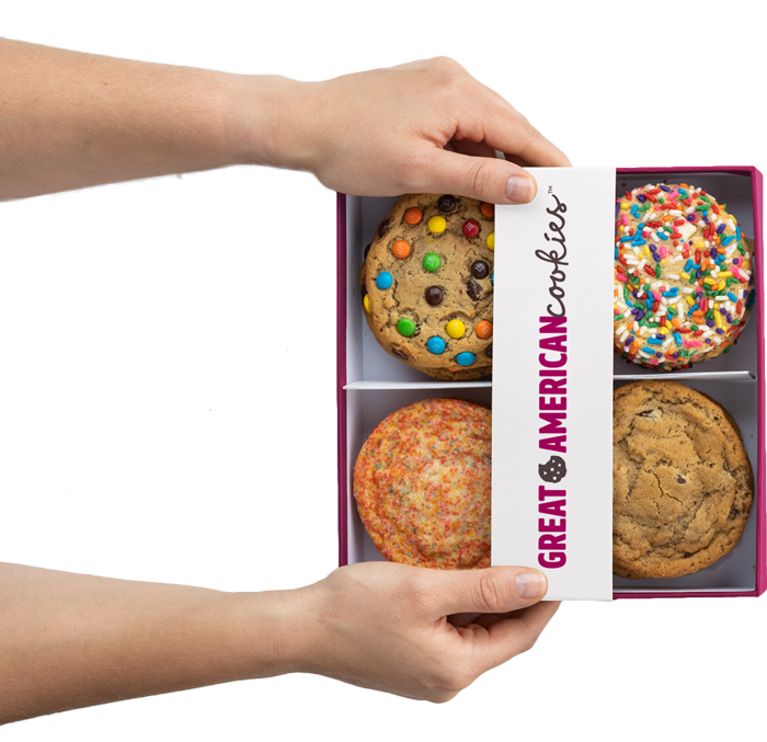 Picture of hands holding a box of assorted cookies including birthday cake, original chocolate chip, m&m and sugar cookies